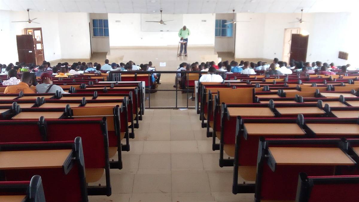 Vast university lecture hall seen from the back rows, a lecturer teaching at the front