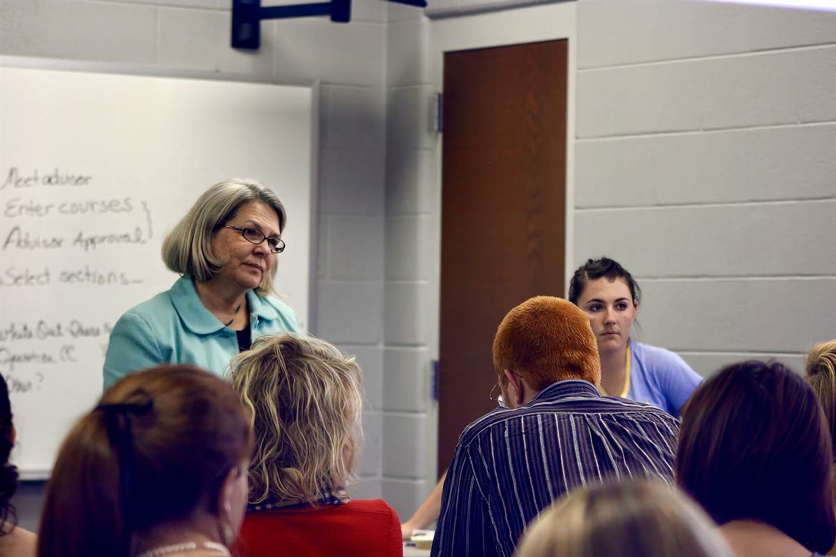 Teacher standing at a whiteboard, listening to a room of adult students seen from behind