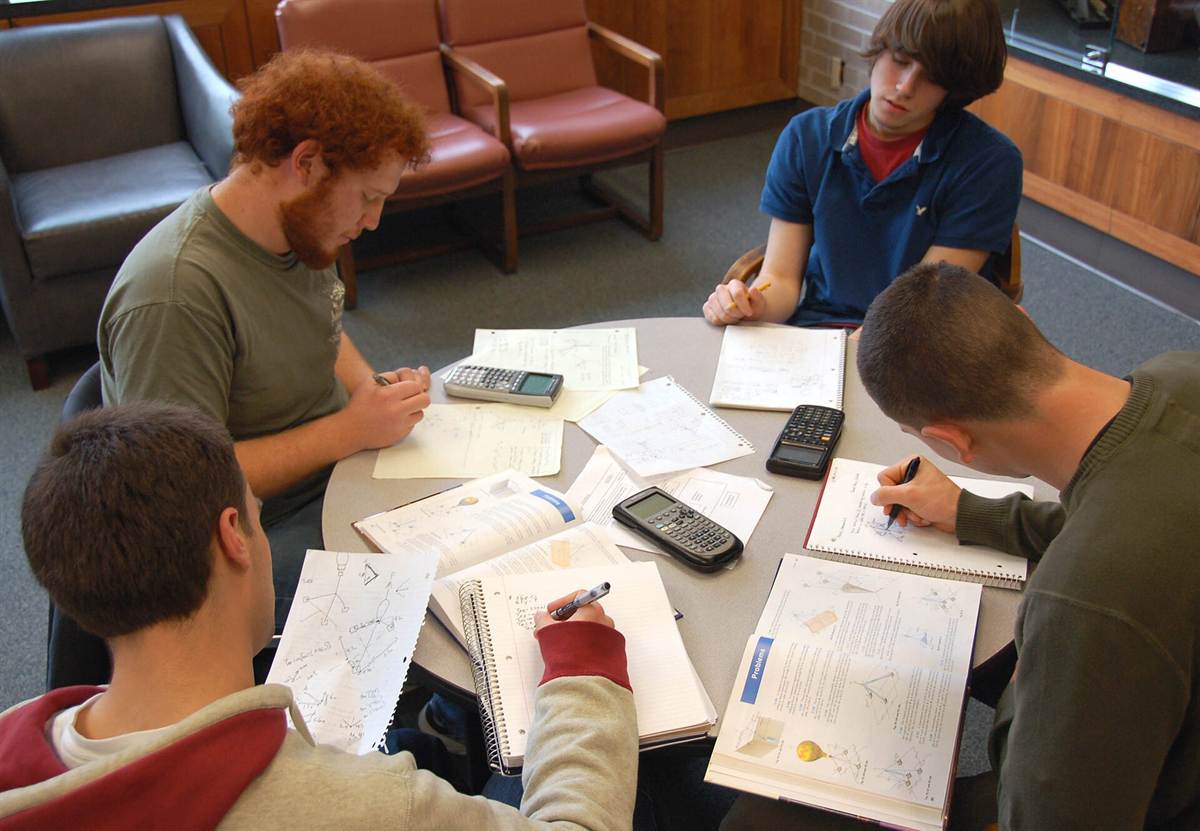 Four students working together around a round table covered with notebooks, textbooks and calculators