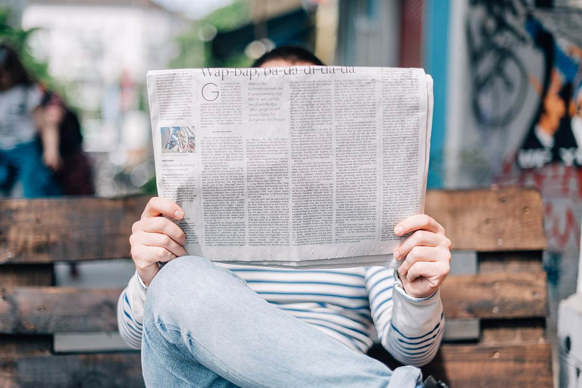 Person sitting on a bench, face hidden behind the wide-open newspaper they are reading